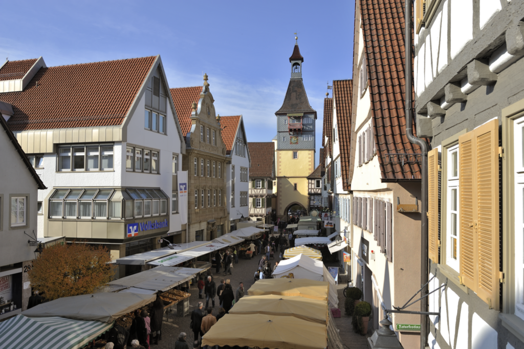 Belebte Marktstraße mit Verkaufsständen, Fachwerkhäusern und historischem Torturm; sichtbar sind die Schriftzüge „Volksbank“, „dm“, „Naturkost“ sowie die Hausnummer „28“.